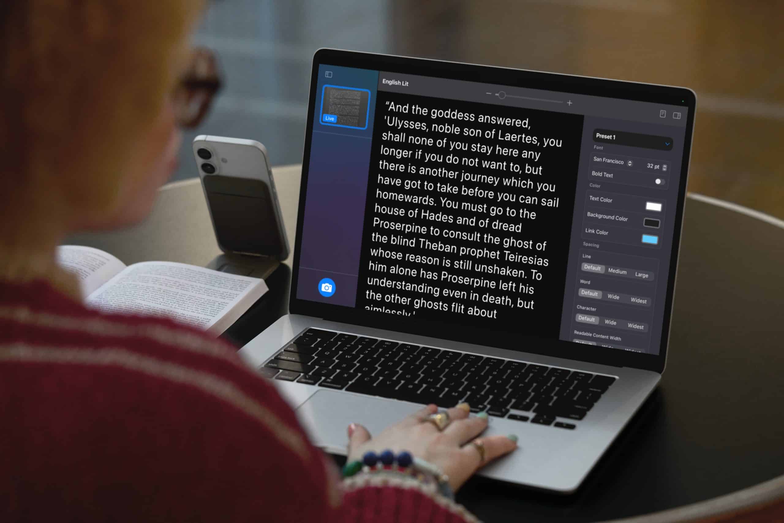 A person sits at a table using a laptop with a text-editing app open, displaying a passage and style options. An open book and a smartphone rest nearby on the table. The person's hands are visible on the laptop keyboard.