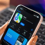 A person holds an iPhone displaying the Apple Maps app page with Apple Maps MICHELIN Ratings featured in the App Store, ready to open, while a laptop is visible in the blurred background.
