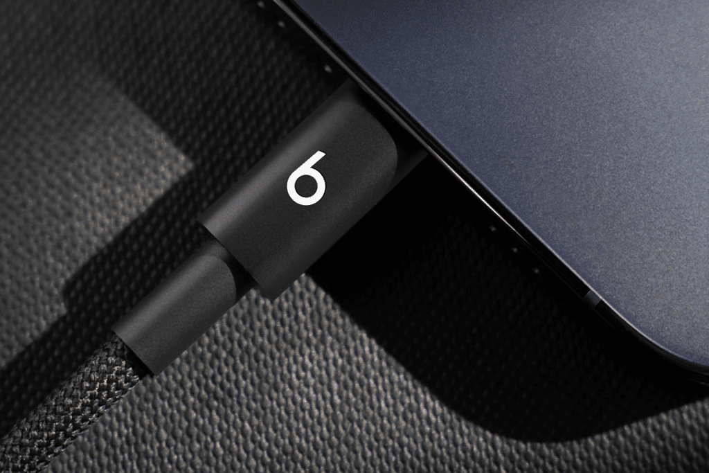 A close-up of a black Beats Cable with a "6" logo plugged into the port of a dark-colored electronic device, possibly a laptop, resting on a textured surface.