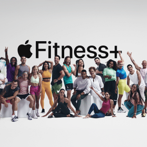 A diverse group of people in athletic wear pose happily together in The Studio, in front of a white background with a large “Apple Fitness+” logo displayed above them.