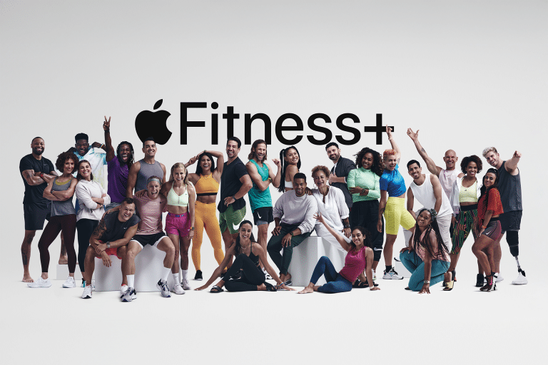 A diverse group of people in athletic wear pose happily together in The Studio, in front of a white background with a large “Apple Fitness+” logo displayed above them.