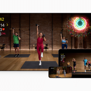 A fitness class from The Studio is displayed on a TV, tablet, phone, and smartwatch, showing three instructors exercising with weights, workout stats, and a celebratory animation in a studio with wooden floors and brick walls.