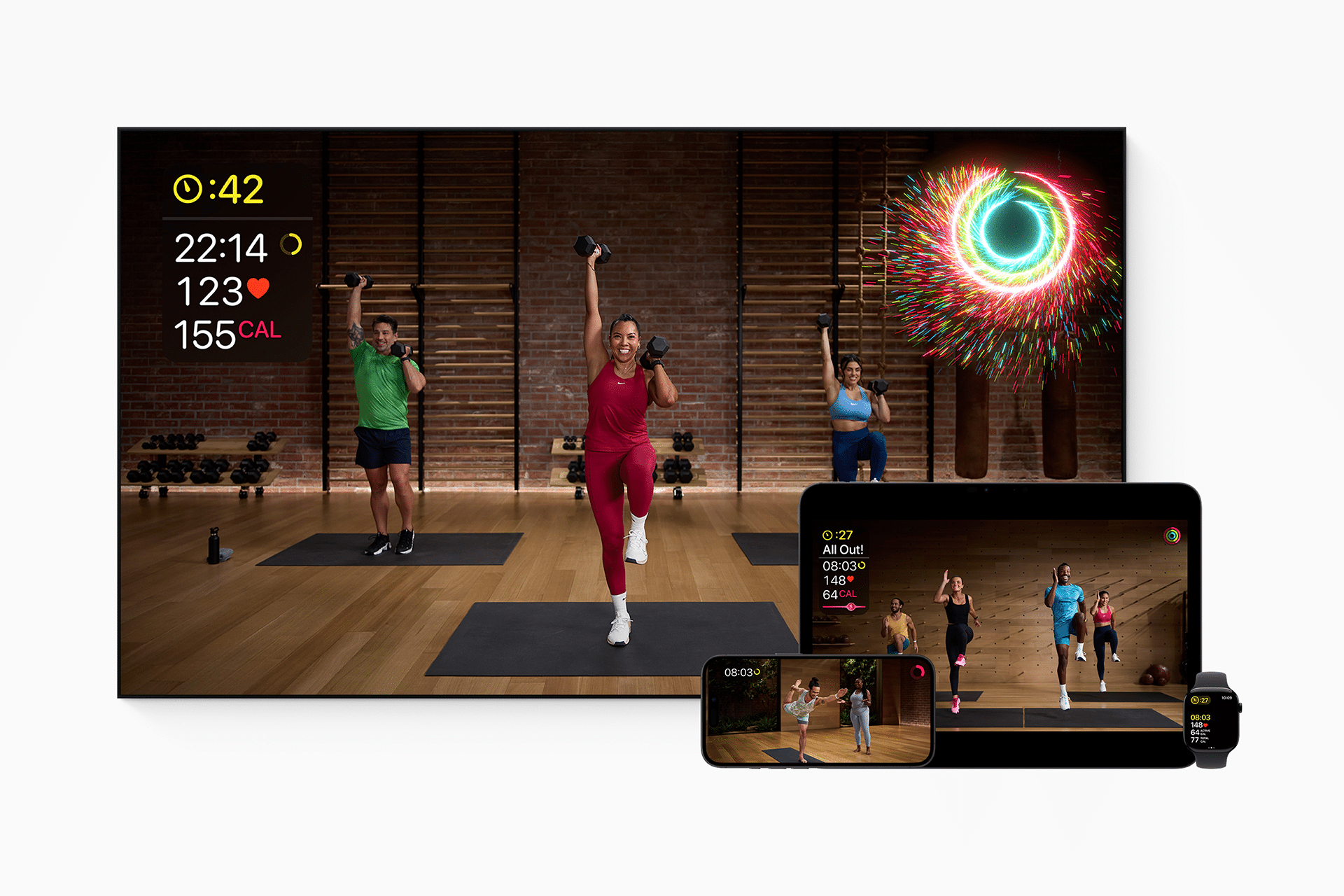 A fitness class from The Studio is displayed on a TV, tablet, phone, and smartwatch, showing three instructors exercising with weights, workout stats, and a celebratory animation in a studio with wooden floors and brick walls.