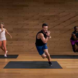 Three people exercise on yoga mats in The Studio’s wooden space, lunging and smiling. The instructor up front wears a black tank top and blue shorts; two participants behind him sport athletic gear in light and dark colors.