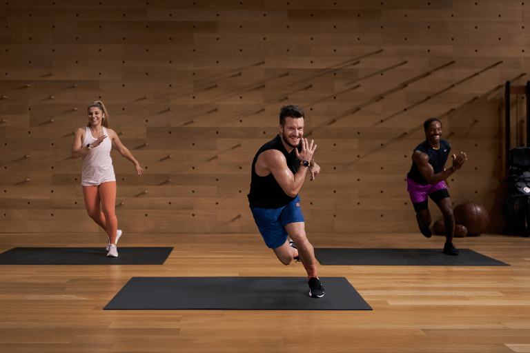 Three people exercise on yoga mats in The Studio’s wooden space, lunging and smiling. The instructor up front wears a black tank top and blue shorts; two participants behind him sport athletic gear in light and dark colors.