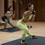 Two women in athletic wear perform squats with dumbbells on black mats at The Studio. With wooden floors, brick walls, and gym equipment in the background, both are focused and smiling as they train together.