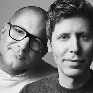 Two men pose closely together in a black-and-white portrait. One wears glasses and a light shirt, resting his head on the other man's shoulder. The second man has short hair and wears a dark top. Both look directly at the camera.