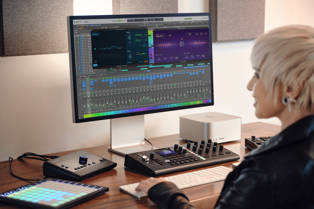 A person with short blonde hair sits at a desk using Logic Pro on a large monitor, surrounded by audio equipment, MIDI controllers, and a computer in a modern studio setting.