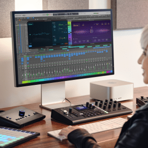 A person with short blonde hair sits at a desk using Logic Pro on a large monitor, surrounded by audio equipment, MIDI controllers, and a computer in a modern studio setting.
