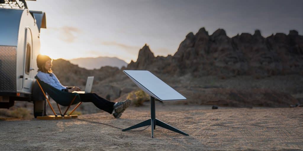 A person with a backpack sits in a chair working on a laptop beside a trailer in a rocky desert landscape at sunset, using Starlink Roam's portable satellite dish for global connectivity.