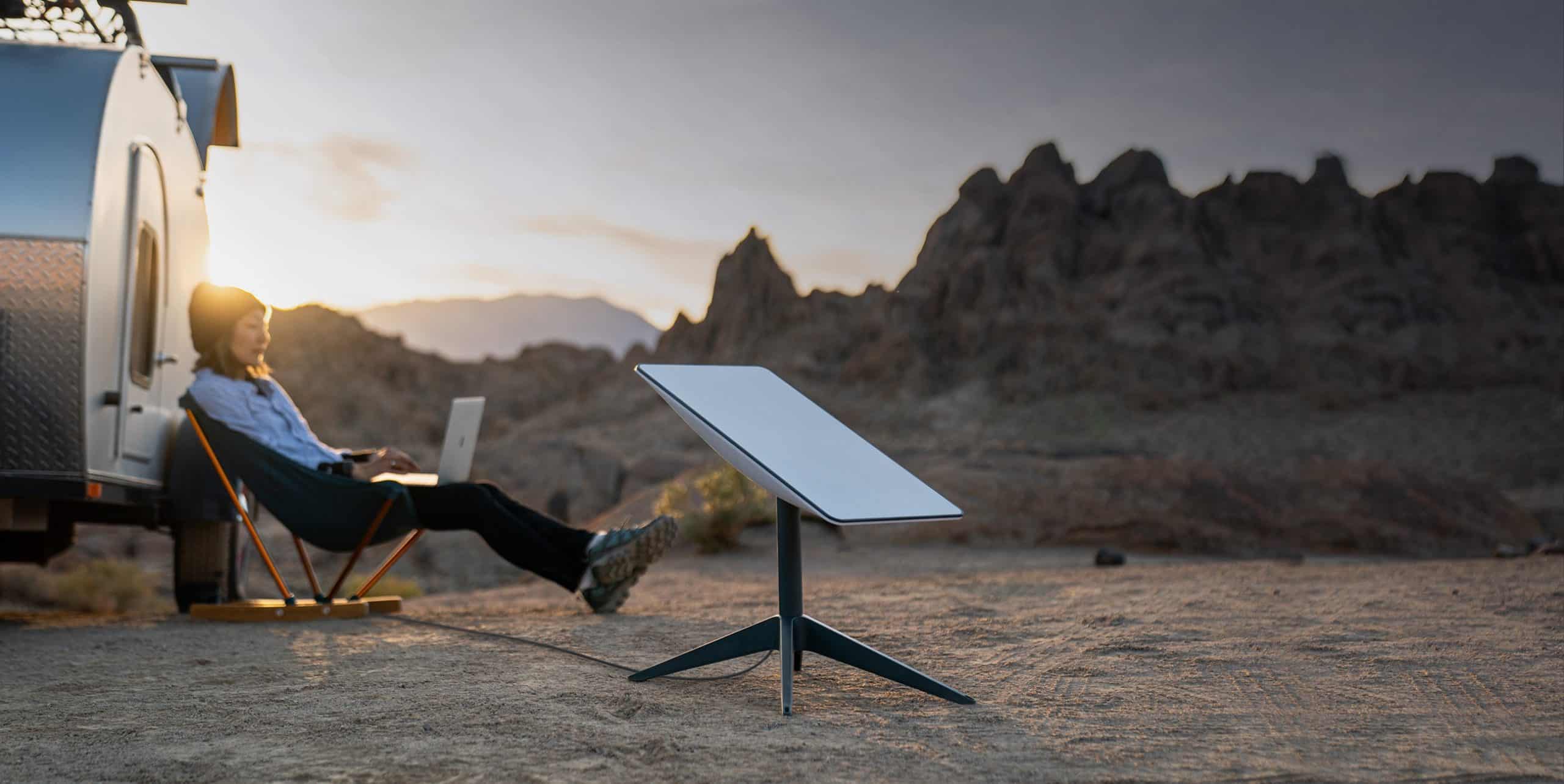 A person with a backpack sits in a chair working on a laptop beside a trailer in a rocky desert landscape at sunset, using Starlink Roam's portable satellite dish for global connectivity.
