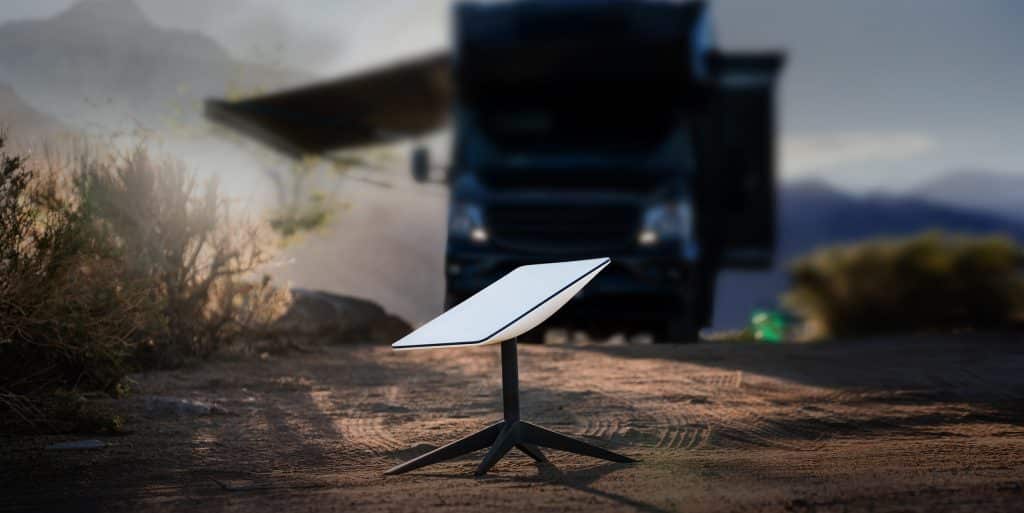 A white Starlink Roam satellite dish on a tripod stand is set up outdoors on a dirt surface, with a blurry RV and mountains in the background, promising global connectivity under soft sunlight.