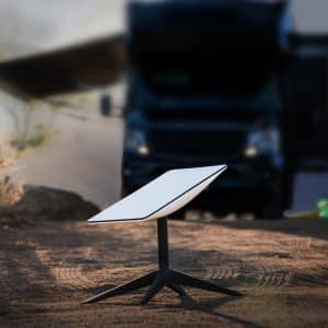 A white Starlink Roam satellite dish on a tripod stand is set up outdoors on a dirt surface, with a blurry RV and mountains in the background, promising global connectivity under soft sunlight.
