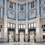 The image shows the exterior of an Apple Store in Den Haag, set within a historic, ornate building featuring tall columns, large windows, and intricate architectural details. The Apple logo is displayed above several entrance doors.
