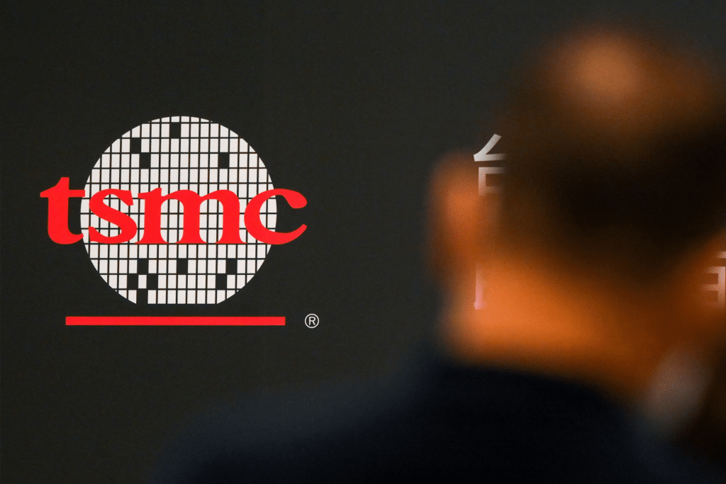 TSMC logo in red letters on a patterned circle, displayed on a dark background. The blurred silhouette of a person appears in the foreground, highlighting TSMC's prominence in the tech industry.