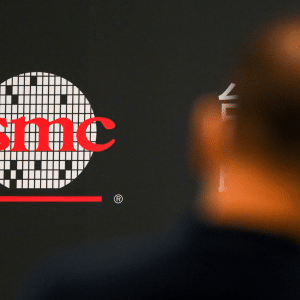 TSMC logo in red letters on a patterned circle, displayed on a dark background. The blurred silhouette of a person appears in the foreground, highlighting TSMC's prominence in the tech industry.