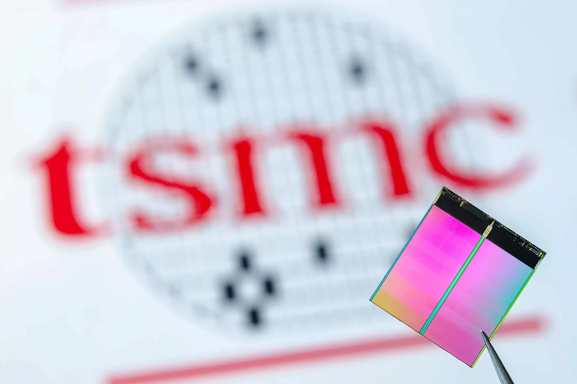A pair of tweezers holds a colorful silicon chip in the foreground, with the blurred TSMC logo in red and black visible in the background.