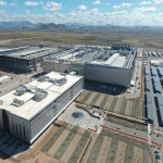 Aerial view of a large, modern TSMC industrial facility with multiple buildings, solar panel arrays, parking lots, and landscaped grounds, set in a desert landscape with mountains in the background under a partly cloudy sky.