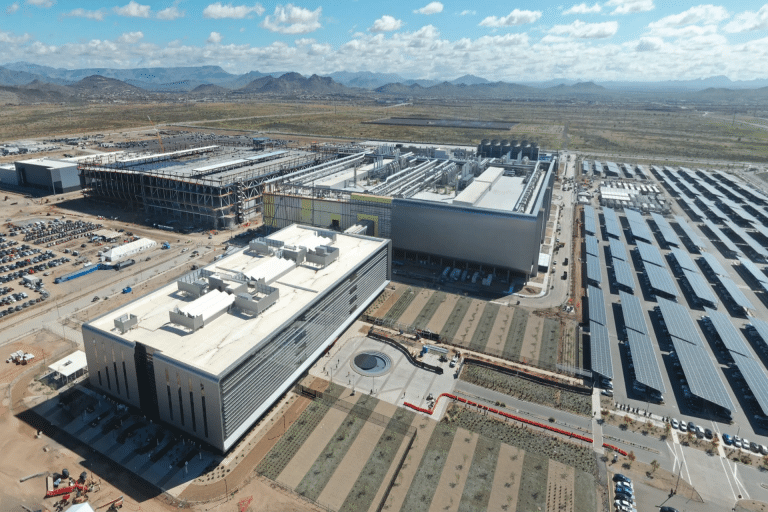 Aerial view of a large, modern TSMC industrial facility with multiple buildings, solar panel arrays, parking lots, and landscaped grounds, set in a desert landscape with mountains in the background under a partly cloudy sky.