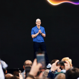A person in a blue shirt and dark pants stands and smiles on stage in front of a large, dark backdrop with a colorful, curved shape above—likely at an Apple Developer App event—as audience members in the foreground hold up phones to take photos.