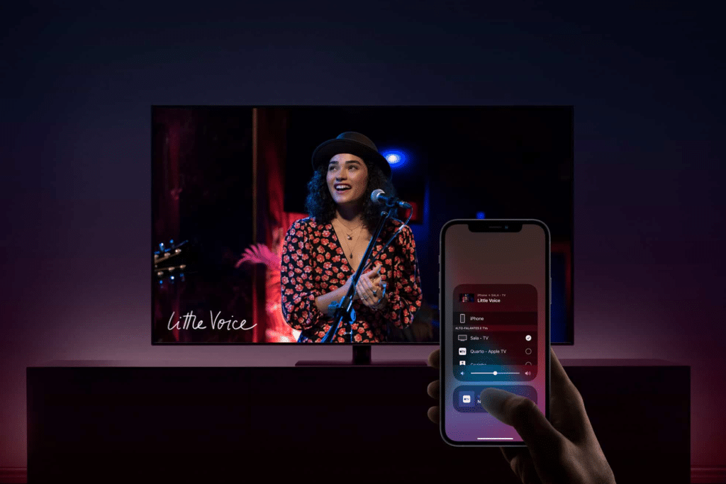 A person uses AirPlay on their smartphone to control media playback while a woman in a hat and red-patterned shirt performs on stage on a TV screen displaying "Little Voice.