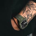 A person with a floral tattoo from modern tattoo studios showcases their personal style, wearing a rugged green Apple Watch on their wrist and resting their hand in a pocket. The Apple logo appears in the lower right corner.