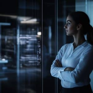 A woman in a white blouse with arms crossed stands in a modern, dimly lit office with glass walls, looking thoughtfully ahead. The Apple logo is visible in the bottom right corner.