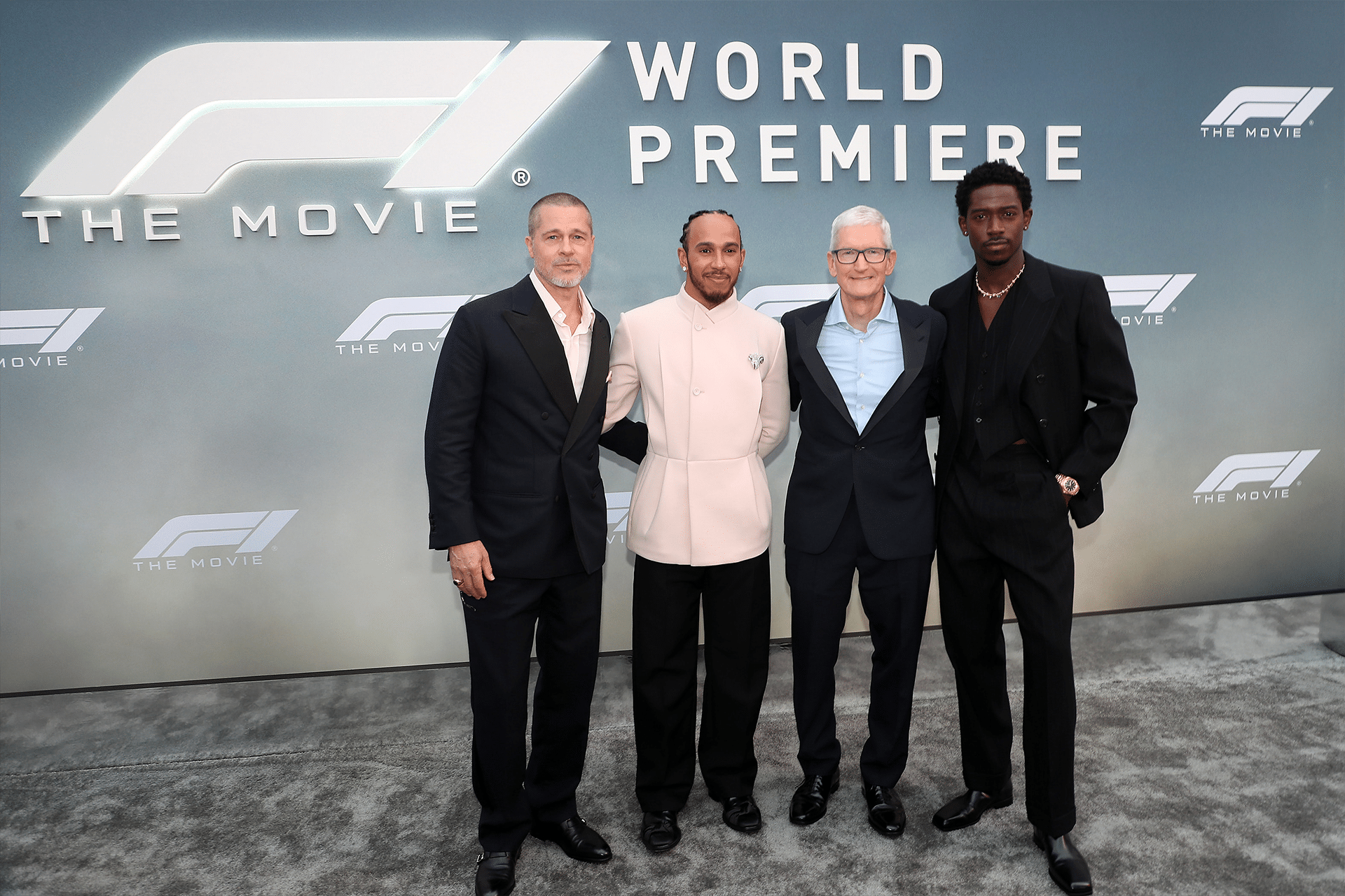 Four men stand together on a grey carpet at the immersive premiere of "F1: The Movie," posing in front of a backdrop with the movie's logo and title. They are dressed in formal and semi-formal attire.