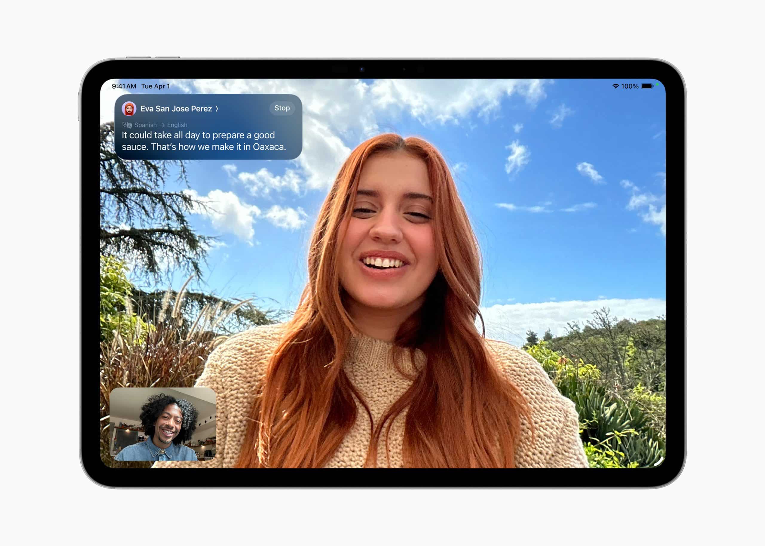 A young woman with long red hair smiles outdoors during a video call on her tablet running iPadOS 26. A message notification appears at the top, and a small window shows another person smiling. Blue sky and greenery fill the background.