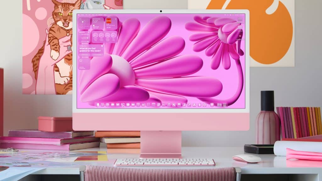 A pink desktop computer running macOS Tahoe with a vibrant pink abstract flower wallpaper sits on a neat desk. Surrounding it are books, art supplies, and colorful wall art, including a cat drawing and orange shapes.