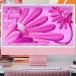 A pink desktop computer running macOS Tahoe with a vibrant pink abstract flower wallpaper sits on a neat desk. Surrounding it are books, art supplies, and colorful wall art, including a cat drawing and orange shapes.
