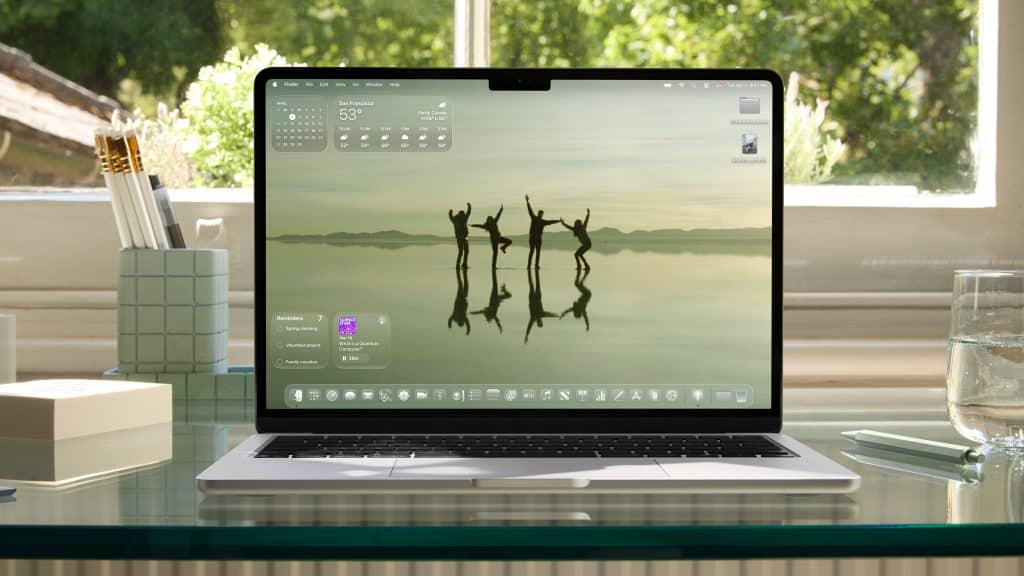 A laptop running macOS Tahoe on a glass desk displays a serene wallpaper of four silhouetted people jumping by a lakeshore. The desktop shows widgets, apps, and weather. The scene is set by a window with trees outside.