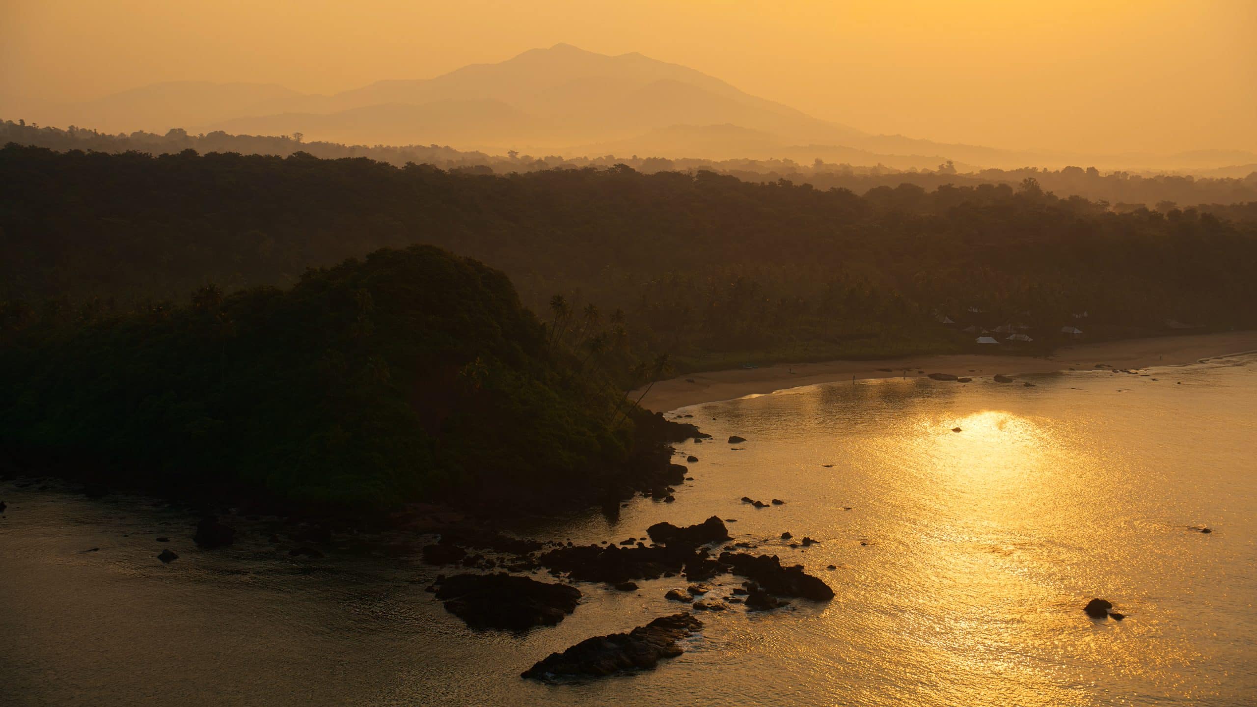 A serene coastal landscape at sunset, with golden sunlight reflecting on the water, lush green hills, scattered rocks, and distant mountains fading into a hazy orange sky—now available as a stunning backdrop on tvOS 26.