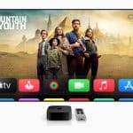 A TV screen running tvOS 26 displays a show titled “Fountain of Youth” with five people standing before ancient-style buildings. Below are colorful app icons, and an Apple TV device with a remote sits in front of the screen.
