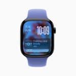 A smartwatch running watchOS 26 with a blue band displays the date "Tue Apr 1" and time "10:09." A notification reads "Training Load Steady -6%" over a blurred background on the screen.