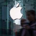 A large white Apple logo is displayed on the glass facade of the Apple Store at La Encantada, with a blurred person holding a drink walking past in the foreground.
