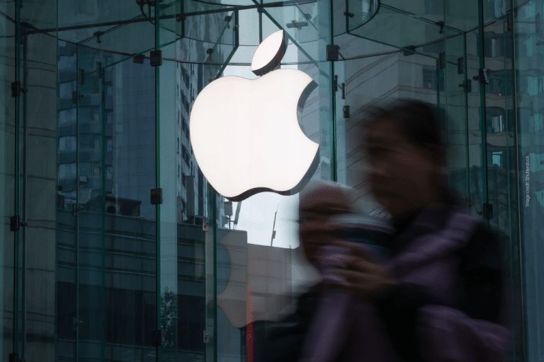 A large white Apple logo is displayed on the glass facade of the Apple Store at La Encantada, with a blurred person holding a drink walking past in the foreground.