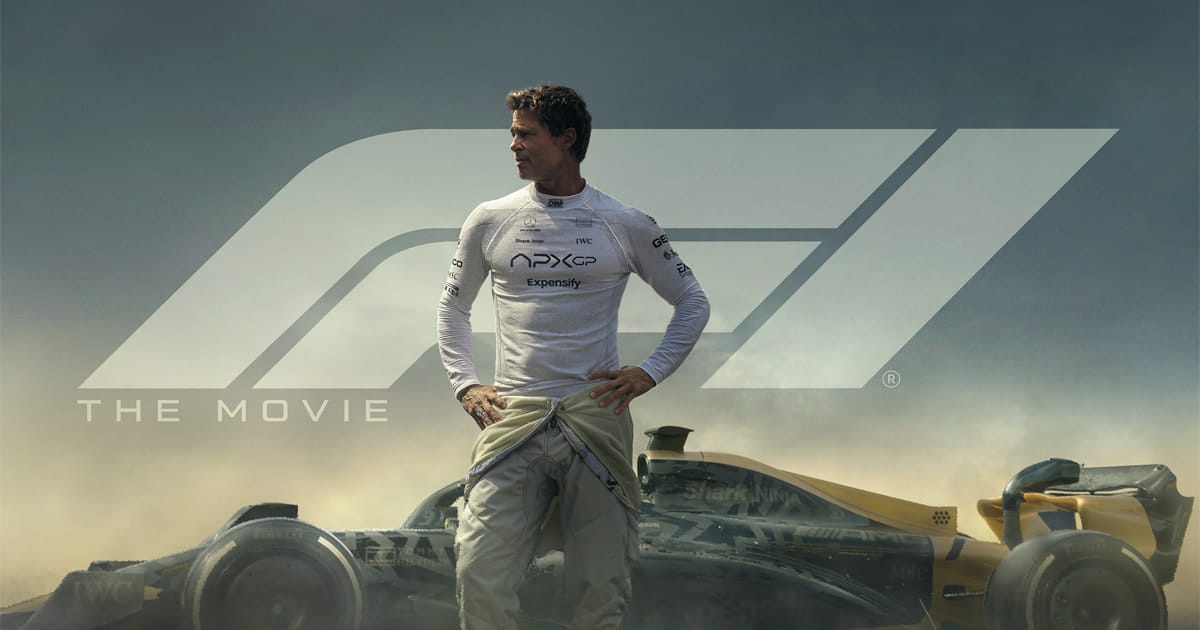 A man in a racing suit stands before a Formula 1 car, hands on hips, looking to the side. The F1 Trailer’s dramatic setting unfolds behind him with a smoky atmosphere and the bold words "THE MOVIE" alongside the iconic F1 logo.
