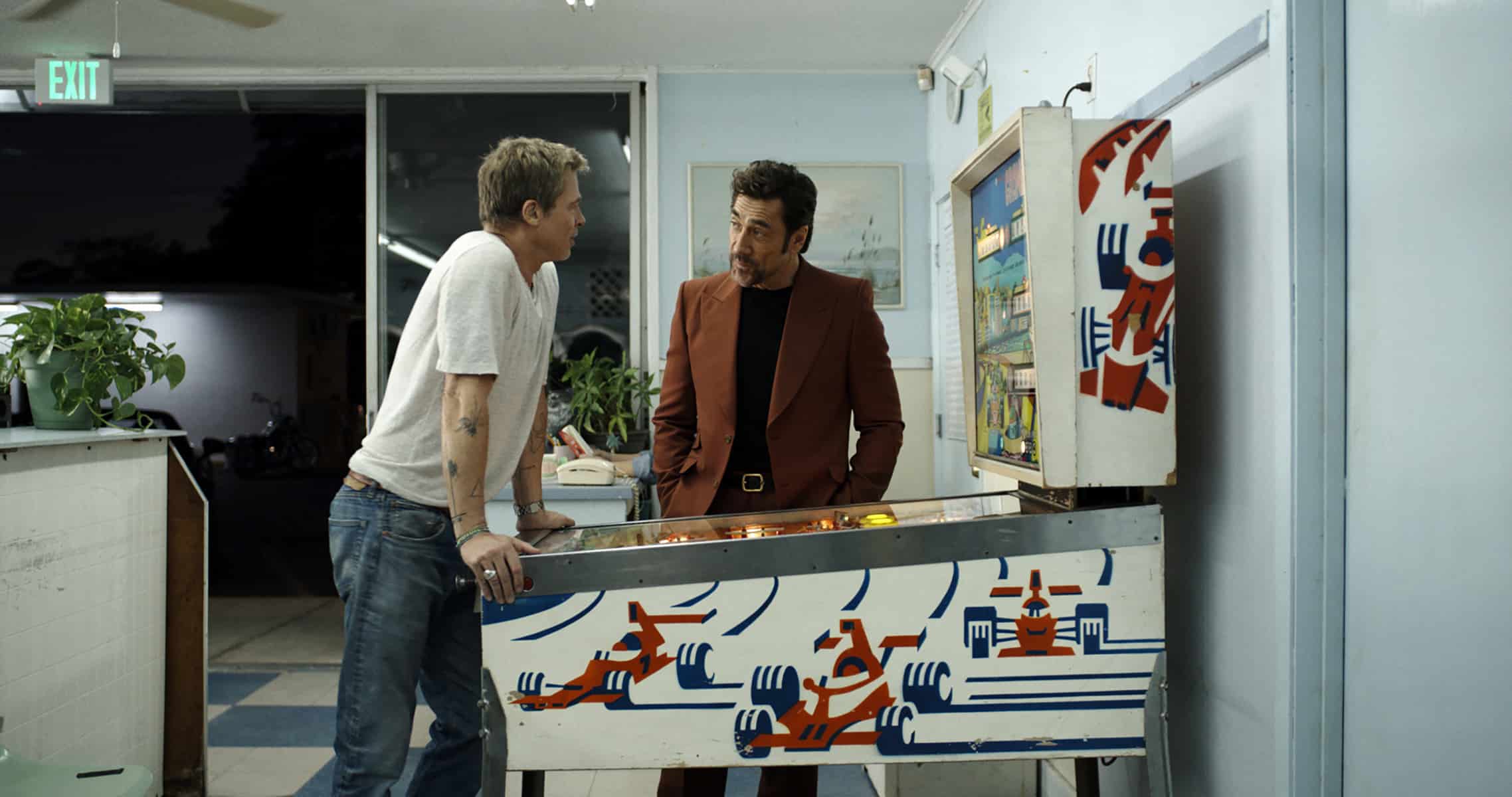 Two men talk by a vintage pinball machine in a room with blue walls; one leans casually in a white t-shirt and jeans, while the other stands upright in a rust-colored suit, as if discussing storytelling for Apple TV+. Plants and a glass door are in the background.