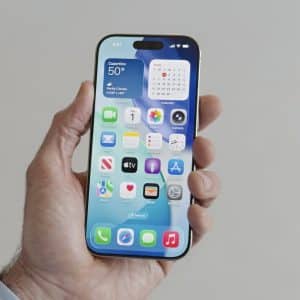 A hand holding an iPhone with a blue wallpaper showcases the Liquid Glass UI on the home screen, featuring various app icons and widgets like weather, calendar, messages, and maps against a neutral background.