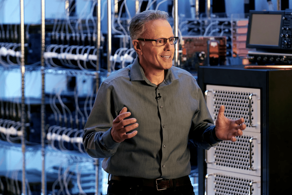 A man wearing glasses and a gray shirt stands in a server room, discussing chip technology as he gestures with his hands. Server racks and electronic equipment are visible in the background.