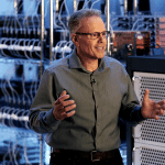 A man wearing glasses and a gray shirt stands in a server room, discussing chip technology as he gestures with his hands. Server racks and electronic equipment are visible in the background.
