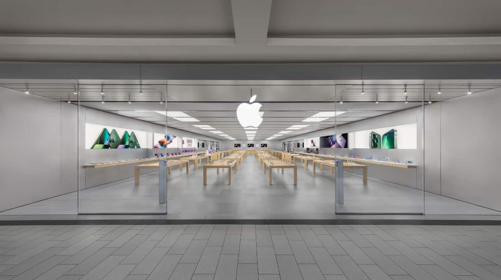 A modern retail space, the Apple Roosevelt Field store features glass walls, wooden tables with electronic devices, and a large white Apple logo above the entrance. The revamped store is clean, bright, organized, and adorned with product images on the walls.