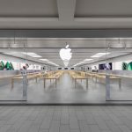 A modern retail space, the Apple Roosevelt Field store features glass walls, wooden tables with electronic devices, and a large white Apple logo above the entrance. The revamped store is clean, bright, organized, and adorned with product images on the walls.
