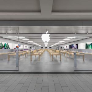 A modern retail space, the Apple Roosevelt Field store features glass walls, wooden tables with electronic devices, and a large white Apple logo above the entrance. The revamped store is clean, bright, organized, and adorned with product images on the walls.