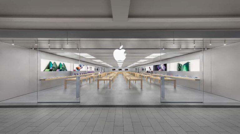 A modern retail space, the Apple Roosevelt Field store features glass walls, wooden tables with electronic devices, and a large white Apple logo above the entrance. The revamped store is clean, bright, organized, and adorned with product images on the walls.