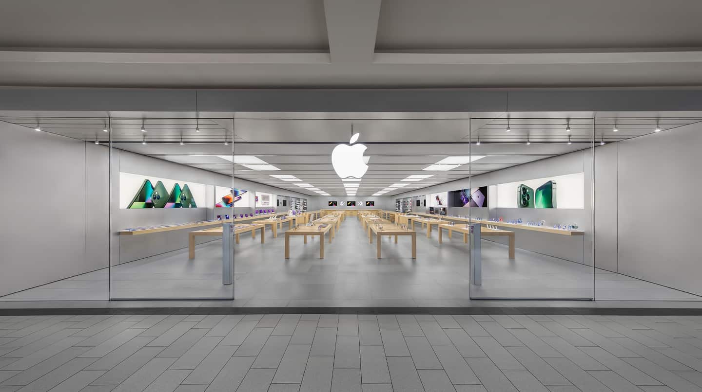 A modern retail space, the Apple Roosevelt Field store features glass walls, wooden tables with electronic devices, and a large white Apple logo above the entrance. The revamped store is clean, bright, organized, and adorned with product images on the walls.