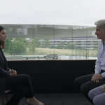 A woman in a dark blazer sits across from two men, one in a blue button-up shirt and the other in a patterned shirt, during a WSJ interview with modern glass buildings and greenery in the background. The WSJ logo appears at the top right.