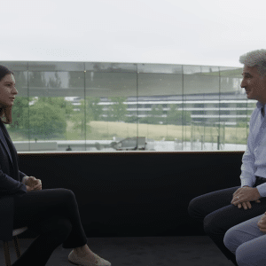 A woman in a dark blazer sits across from two men, one in a blue button-up shirt and the other in a patterned shirt, during a WSJ interview with modern glass buildings and greenery in the background. The WSJ logo appears at the top right.