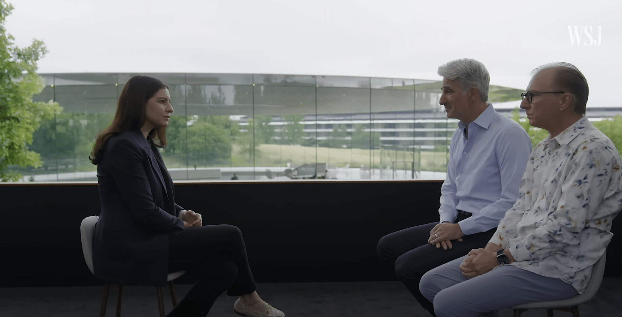 A woman in a dark blazer sits across from two men, one in a blue button-up shirt and the other in a patterned shirt, during a WSJ interview with modern glass buildings and greenery in the background. The WSJ logo appears at the top right.